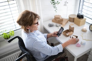 A disabled mature woman in wheelchair packing handmade products indoors at home, small business concept.