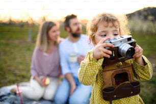 Small girl with camera taking photograph of family on picnic outdoors in spring nature at sunset.