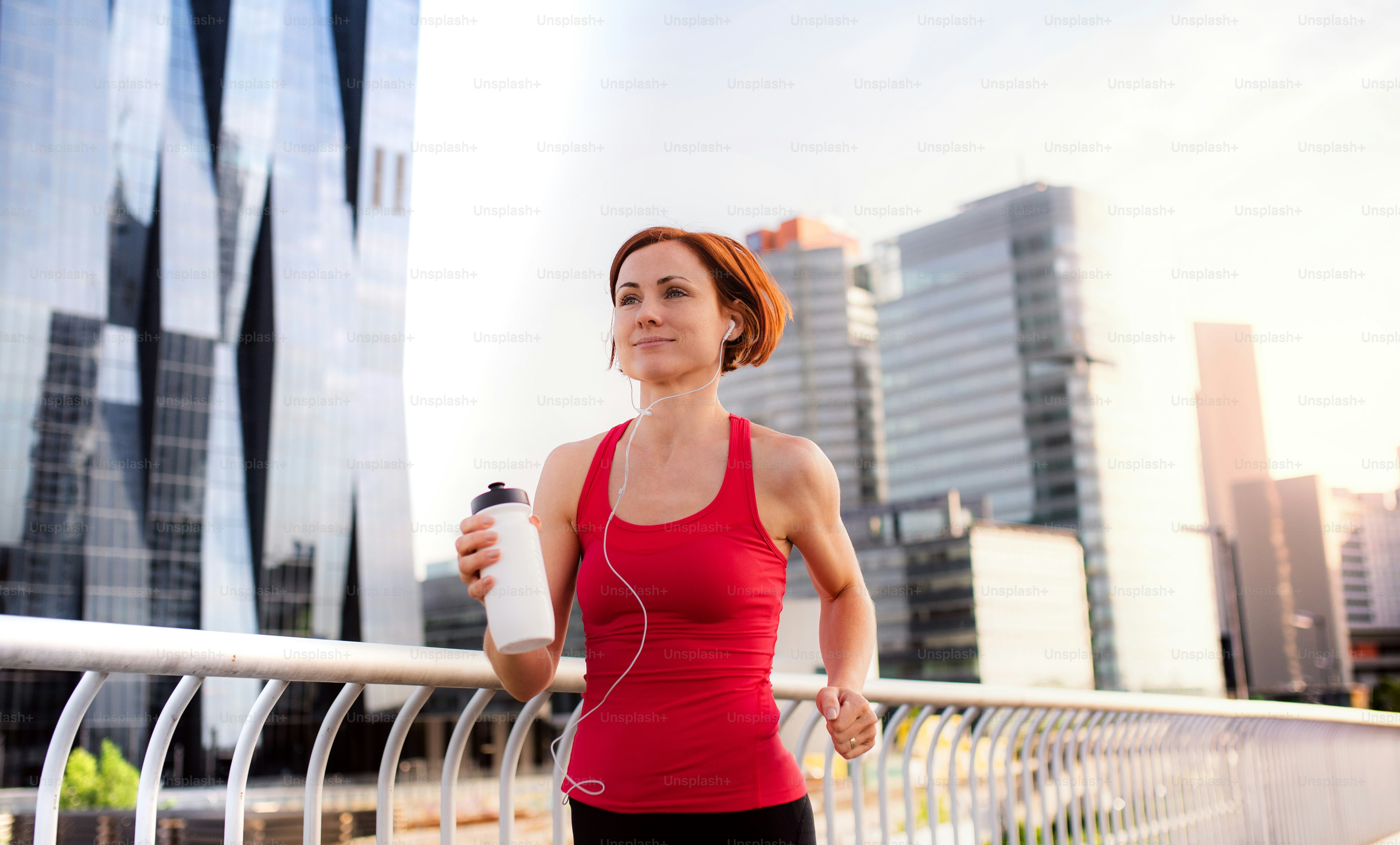 Active young woman runner with earphones and water bottle jogging outdoors in city.