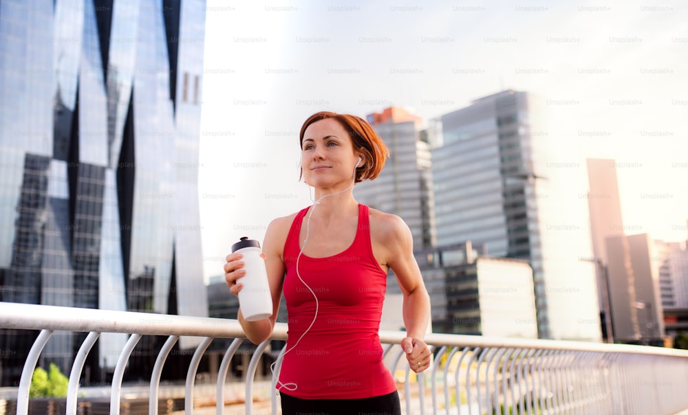 Active young woman runner with earphones and water bottle jogging outdoors in city.