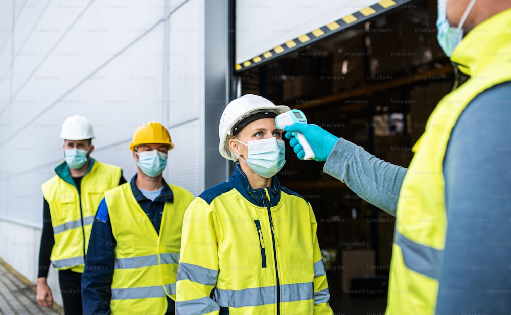 Gruppo di lavoratori con maschera facciale in piedi davanti al magazzino, al coronavirus e al concetto di misurazione della temperatura.