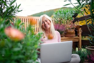 A senior woman with laptop sitting outdoors on terrace, working.
