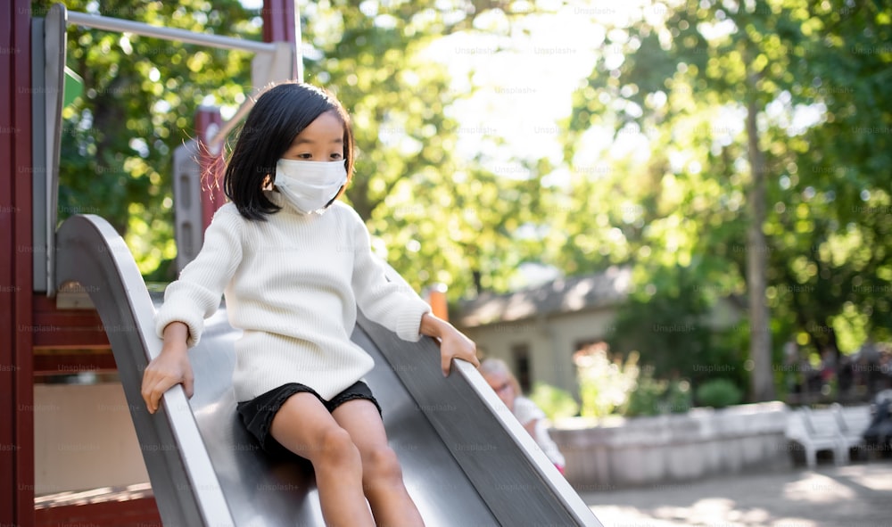 Small girl with face mask on slide on playground outdoors in town, coronavirus concept.