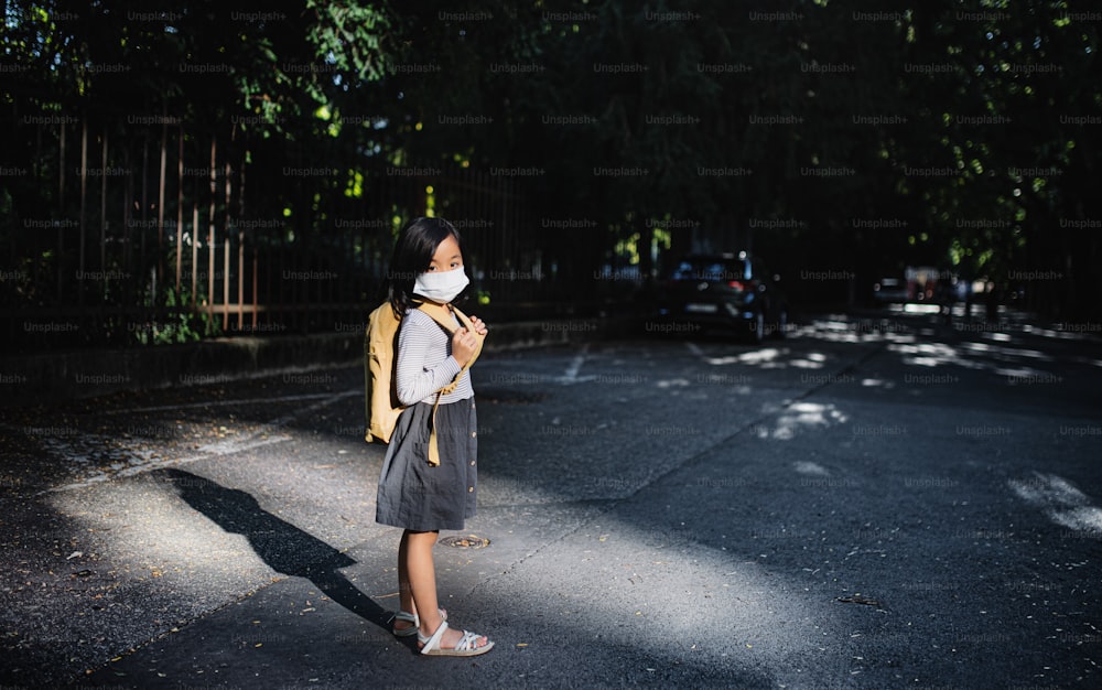 Portrait of small Japanese girl with backpack standing outdoors in town, coronavirus concept. Copy space.