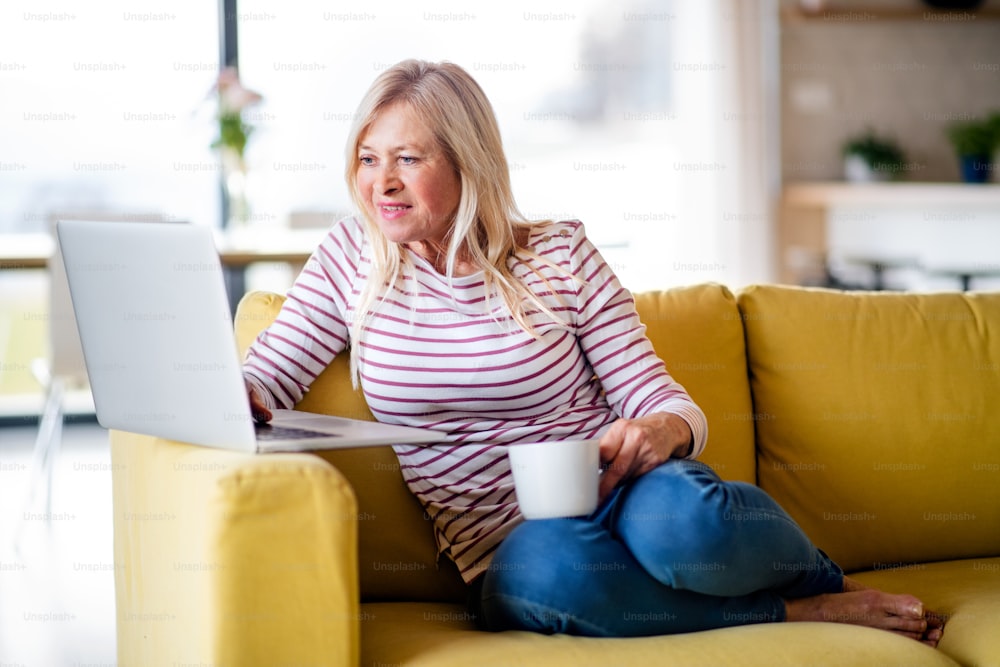 Mulher sênior com café e laptop dentro de casa, relaxando no sofá.
