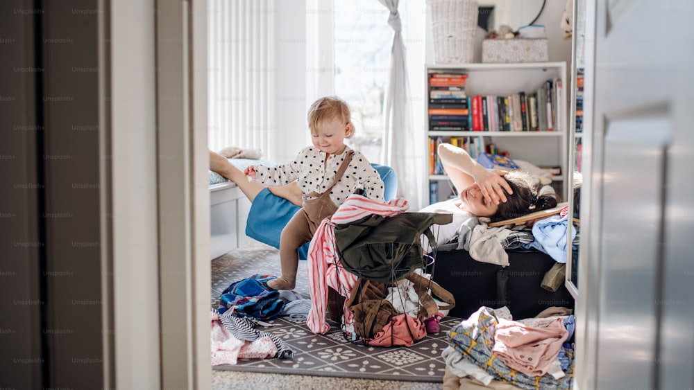 Tired and frustrated mother with small toddler daughter in messy bedroom at home, resting.