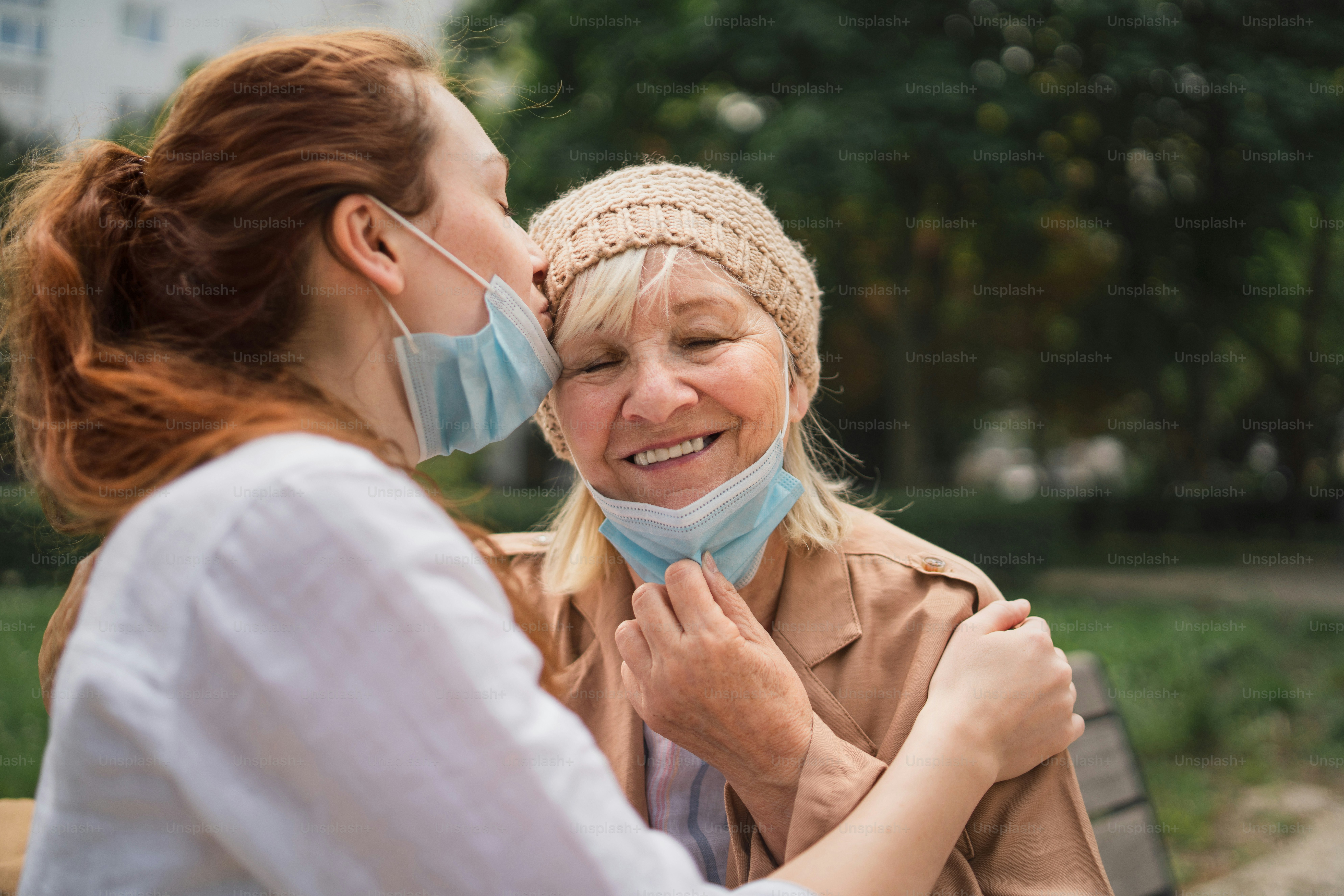A portrait of young woman kissing grandmother outdoors in city, life after covid-19 vaccination.