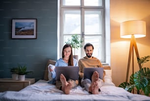 Vista frontal de una pareja joven que usa computadoras portátiles en la cama en el interior, concepto de oficina en casa.