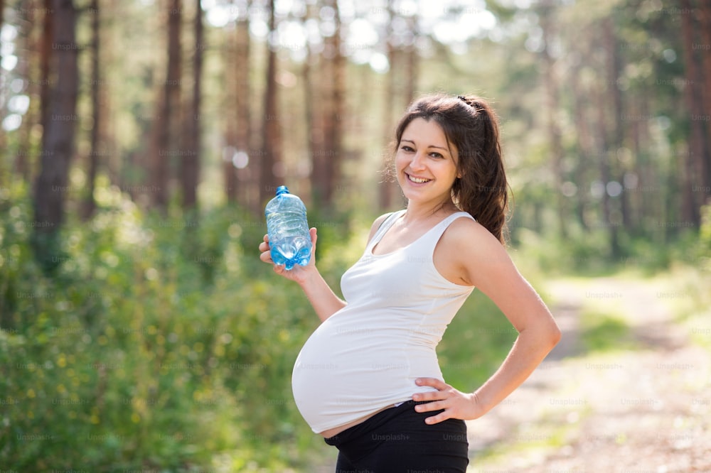 Pregnant woman outdoors in nature, holding water bottle after exercise and looking at camera.