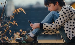A side view of preteen girl with smartphone sitting outdoors in town.