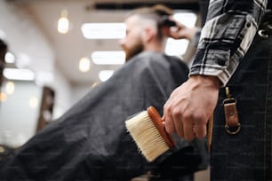 Jeune homme client dans un salon de coiffure, barbier méconnaissable tenant une brosse.