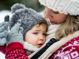 Gros plan d’une petite fille bouleversée dans un porte-bébé et d’une mère méconnaissable dans la nature hivernale.