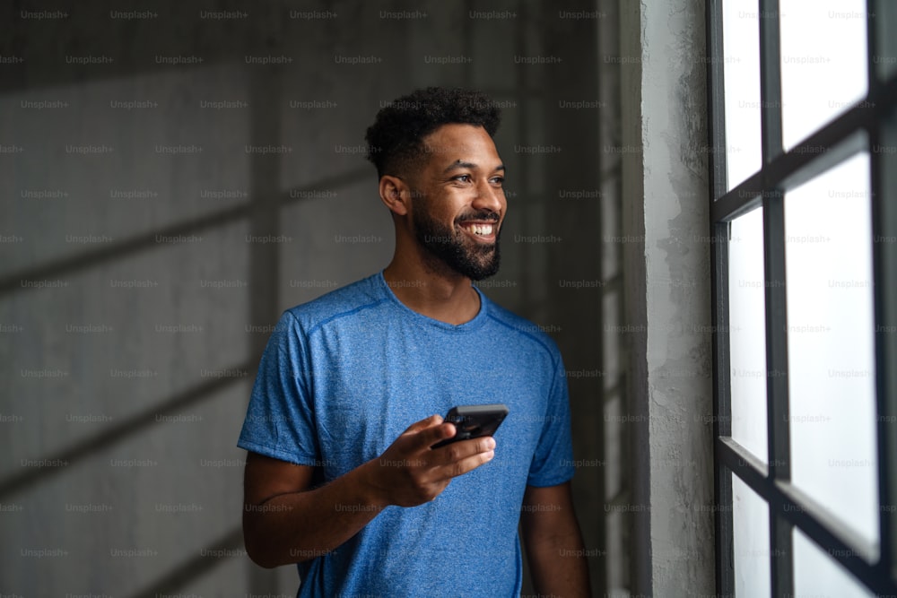 A happy young African American sportsman standing indoors at gym, using smartphone.