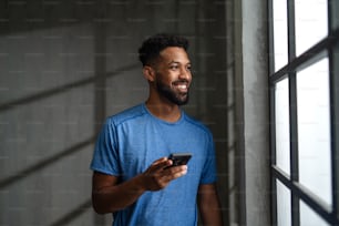 Un joven deportista afroamericano feliz parado en el interior del gimnasio, usando un teléfono inteligente.