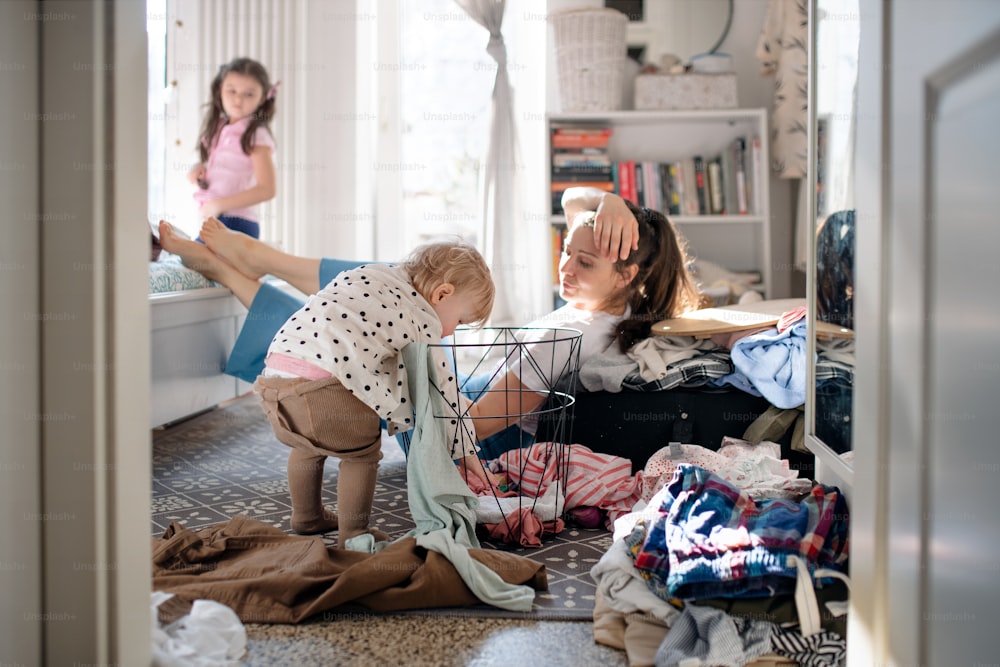 Tired and frustrated mother with small toddler daughter in messy bedroom at home, resting.