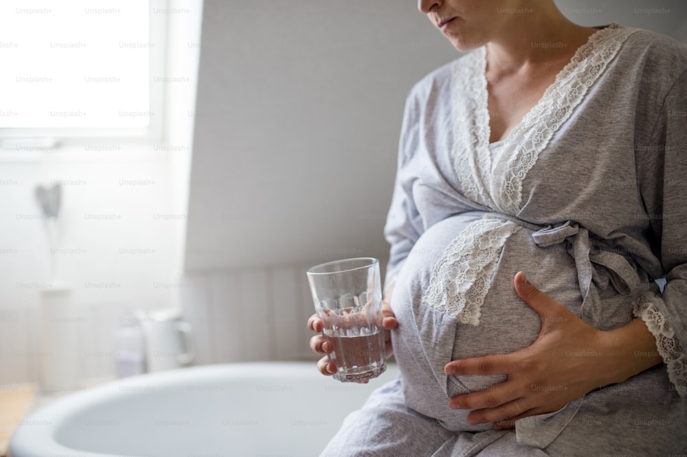 Seção média de mulher grávida irreconhecível dentro de casa no banheiro em casa, segurando copo de água.