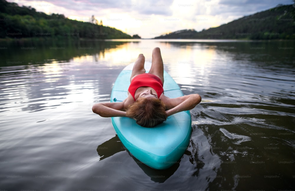 Vista posteriore della donna anziana contenta sdraiata sul paddleboard sul lago in estate, a riposo.