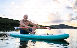 Front view of senior man sitting on paddleboard on lake in summer. Copy space.
