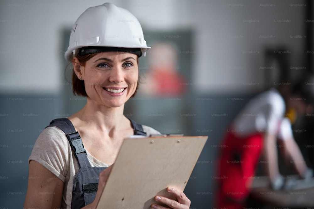 A portrait of woman worker with helmet indoors in factory holding clipboard.