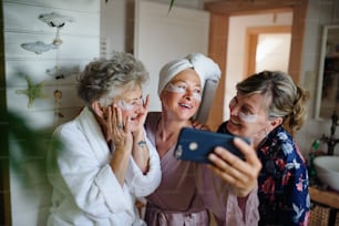Happy senior women friends in bathrobes taking a selfie indoors at home, selfcare concept.