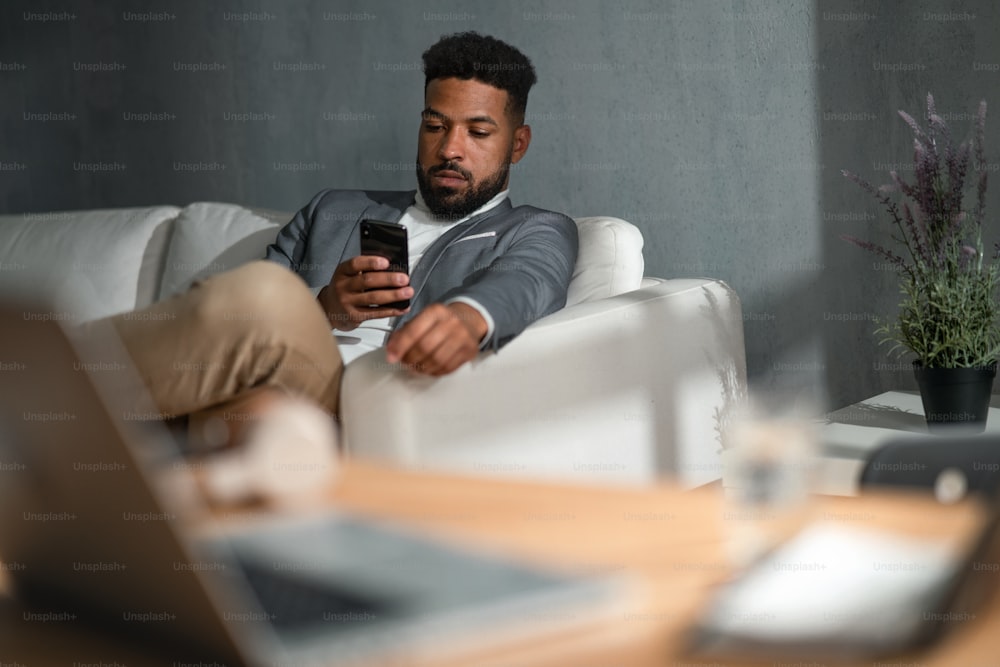 A young african american businessman with smartphone sitting on sofa indoors in office