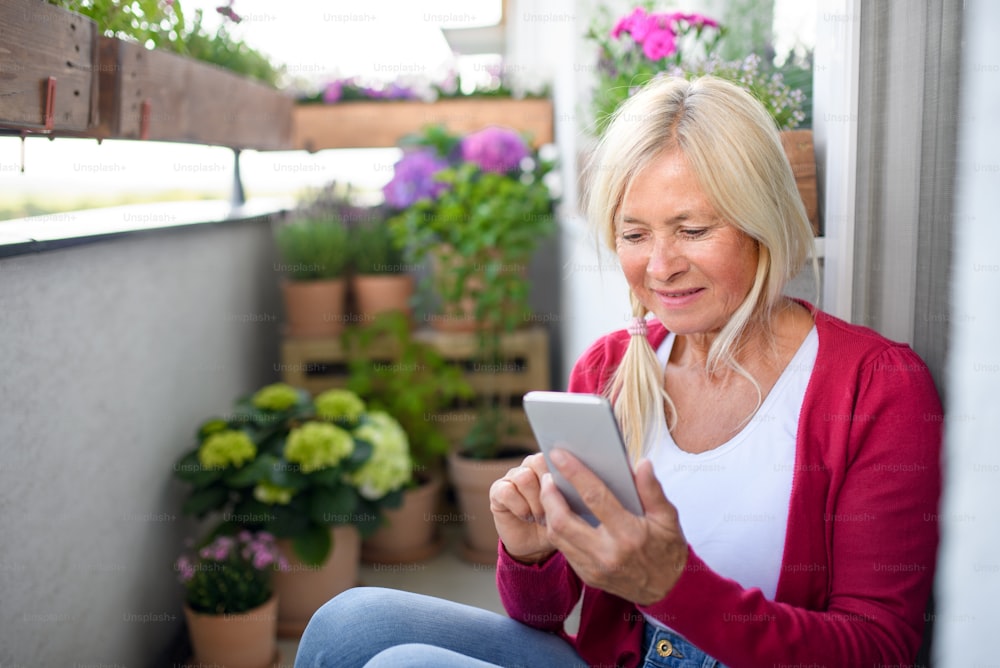 Happy senior woman relaxing on balcony in summer, using smartphone.