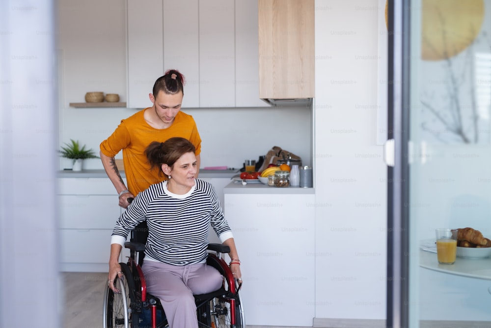 Un figlio adulto che aiuta la madre matura disabile in sedia a rotelle in cucina all'interno di casa.
