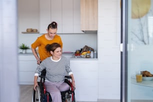 An adult son helping disabled mature mother in wheelchair in kitchen indoors at home.