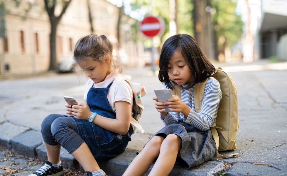 De petites filles avec des smartphones assises à l’extérieur en ville, jouant à des jeux en ligne.