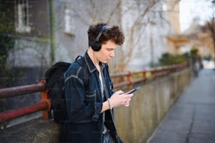 A portrait of young man commuter with headphones standing outdoors in city, using smartphone.