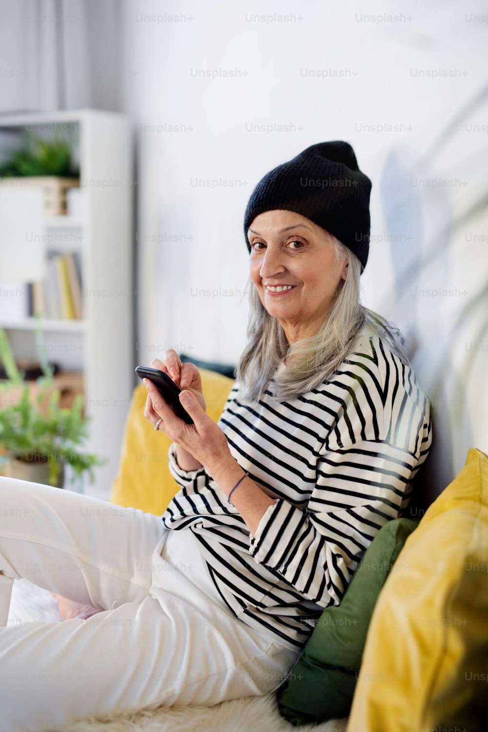 Retrato da mulher idosa com chapéu sentado dentro de casa em casa, usando o smartphone.