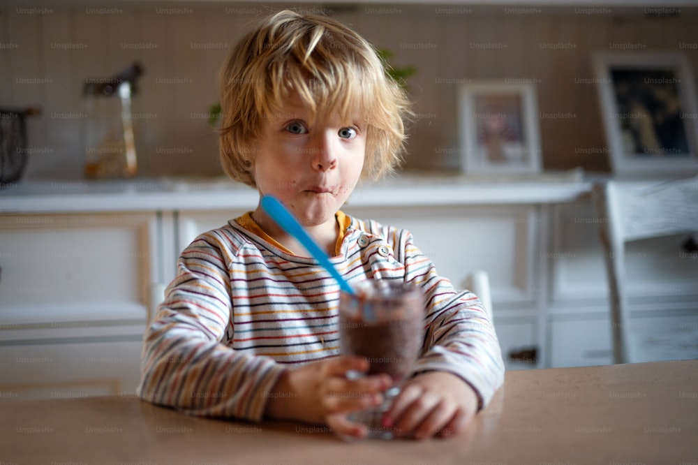 Porträt eines kleinen Jungen mit schmutzigem Mund drinnen in der Küche zu Hause, der Pudding isst.