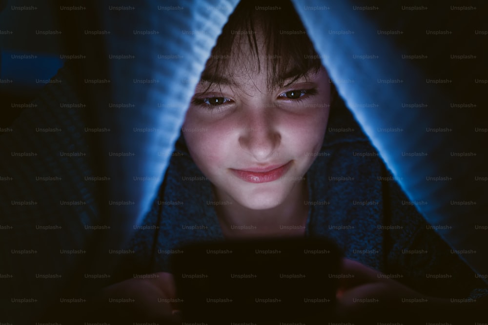 A happy teen girl using smartphone, hiding under blanket at nigh, social networks cocnept.