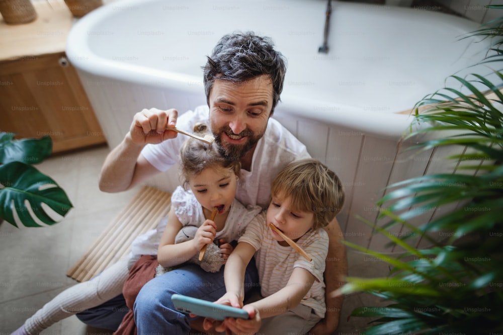 Visão de alto ângulo de um pai com duas crianças pequenas escovando os dentes dentro de casa e tirando selfie, conceito de estilo de vida sustentável.