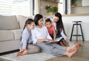Portrait de petites filles heureuses avec leur mère et leur grand-mère à l’intérieur de la maison, lisant un livre d’histoires.
