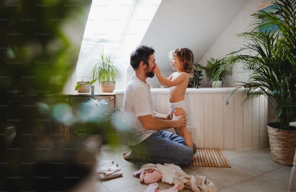 Side view of mature father with small daughter indoors at home, getting ready for a bath.