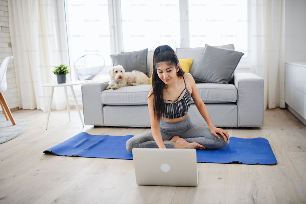 Un retrato de una mujer joven del deporte con una computadora portátil haciendo ejercicio en el interior de la casa, concepto de estilo de vida saludable.