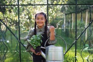 A happy small girl gardening in greenhouse outdoors in backyard, looking at camera.