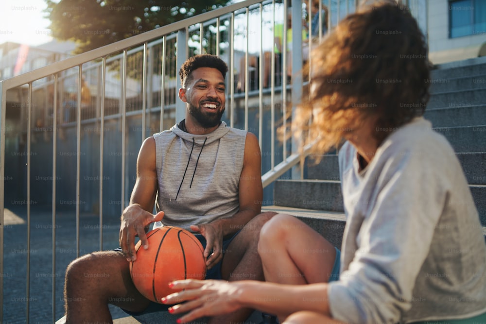 A man and woman friends with basketball resting after exercise outdoors in city, talking.