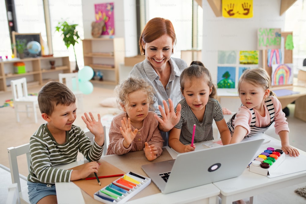Un grupo de pequeños niños de guardería con maestra en el interior del aula, usando una computadora portátil.