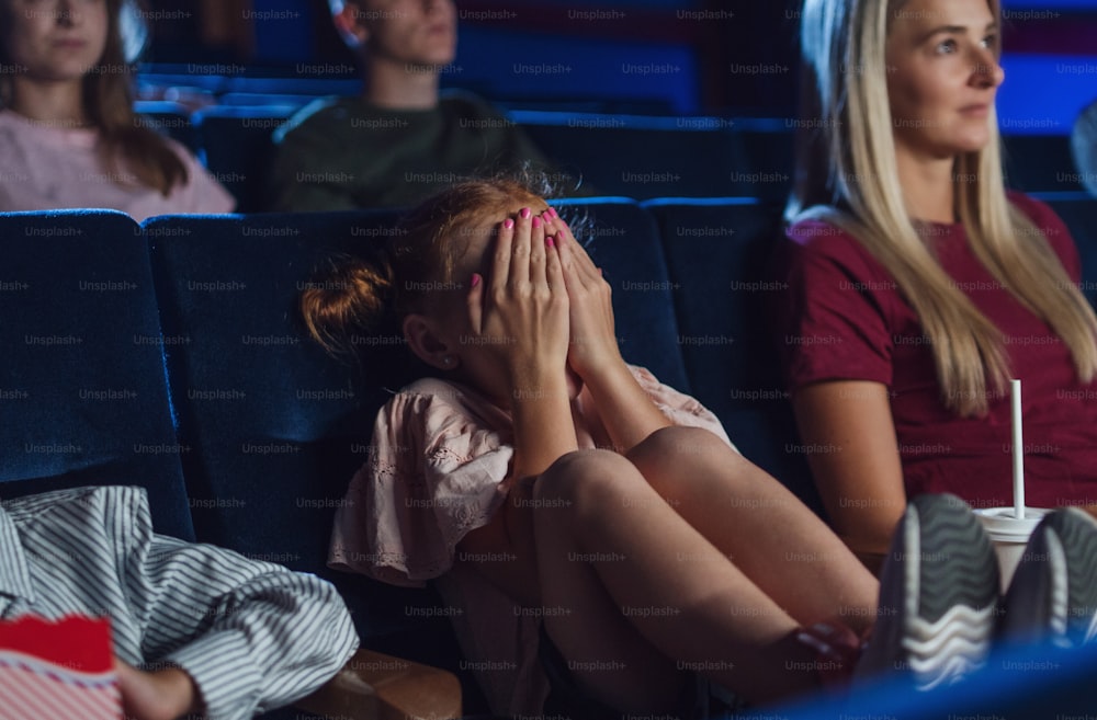A worried small girl with mother covering eyes in the cinema while watching film.