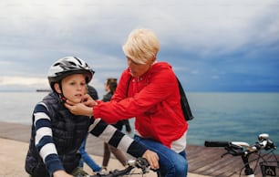 Mother and small son with bicycles outdoors standing on beach, putting on helmet.