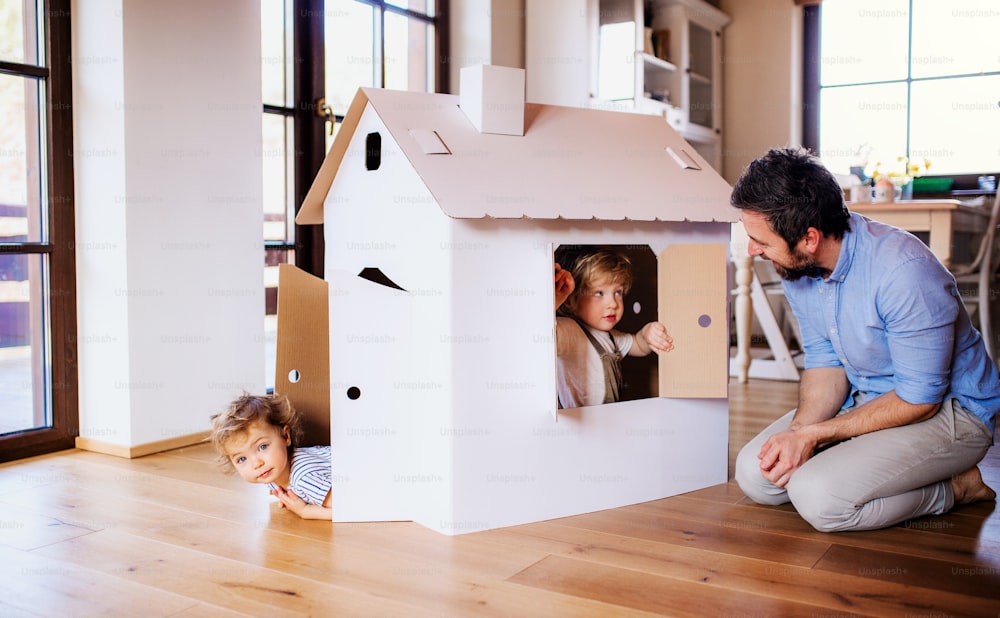 Two happy toddler children with father playing with paper house indoors at home.