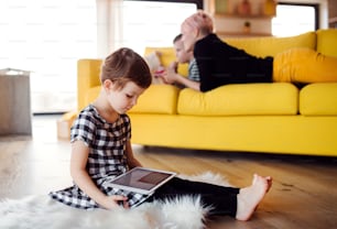 A small girl with mother and brother using tablet indoors on the floor at home.