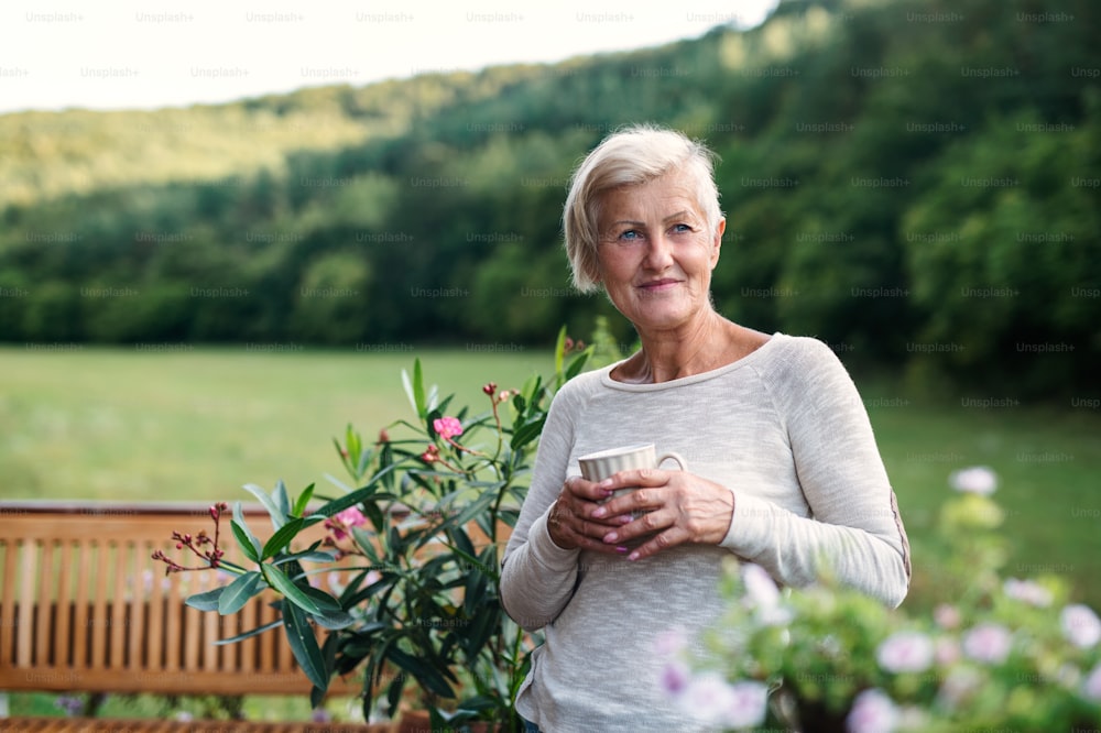 A senior woman with coffee standing outdoors on a terrace in summer.