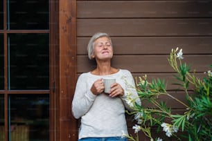 Eine ältere Frau mit Kaffee steht im Sommer draußen auf einer Terrasse, die Augen geschlossen.