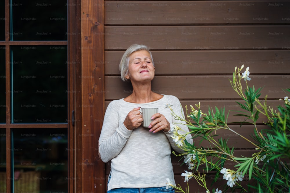 Una donna anziana con il caffè in piedi all'aperto su una terrazza in estate, gli occhi chiusi.