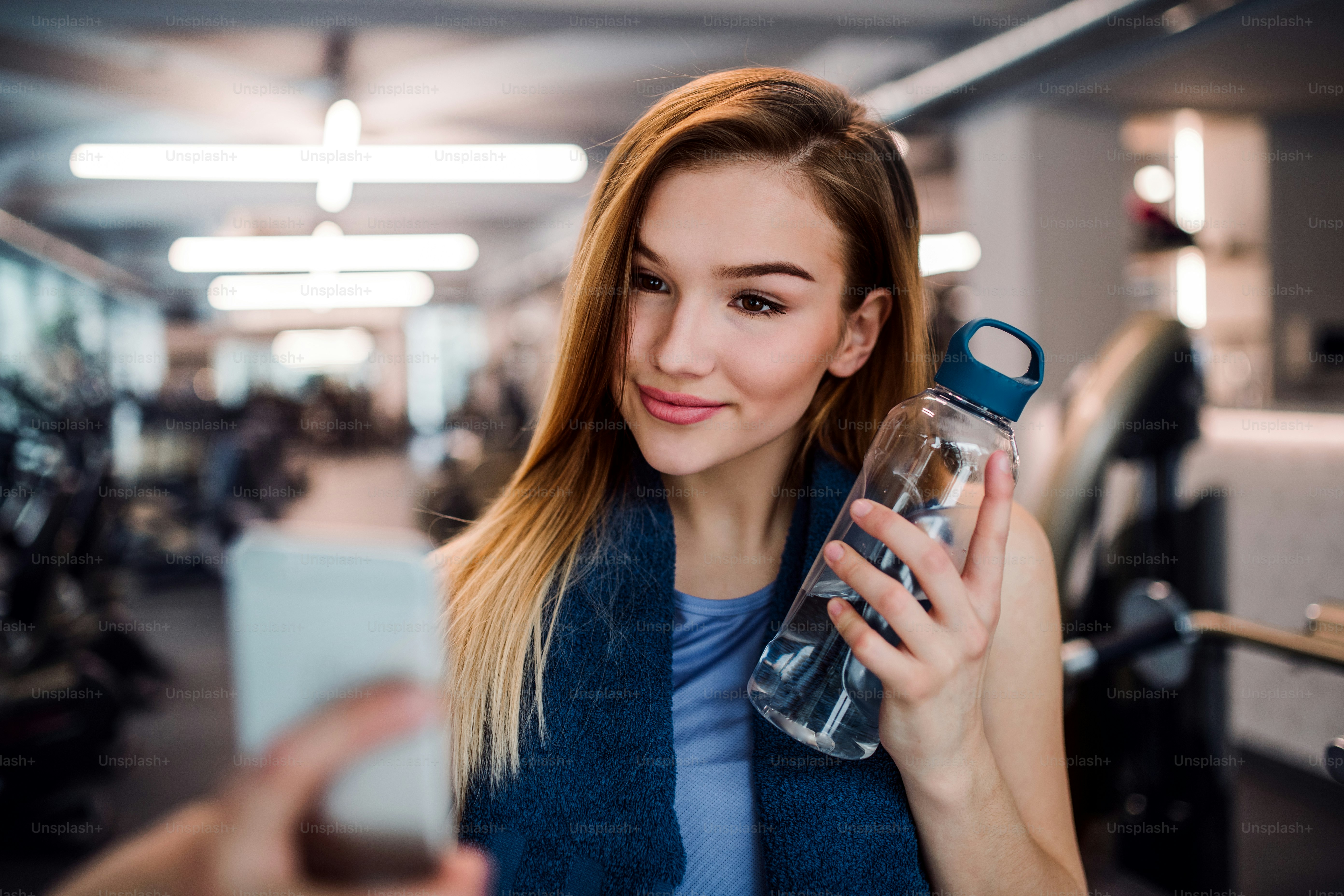 Un retrato de una niña o mujer joven con una botella de agua y un teléfono inteligente en un gimnasio, tomando una selfie.