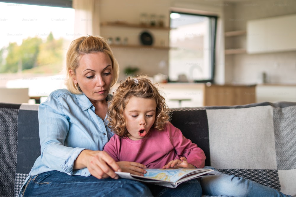 Una bambina carina con la madre seduta sul divano all'interno di casa, leggendo un libro di storie.