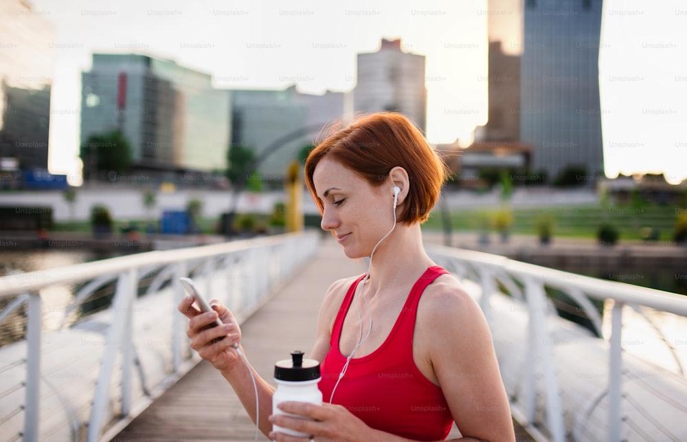A young woman resting after doing exercise on bridge outdoors in city, using smartphone.
